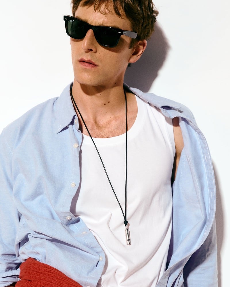 Quentin Demeester in a light blue oxford shirt worn open over a white tank top with a whistle necklace and sunglasses