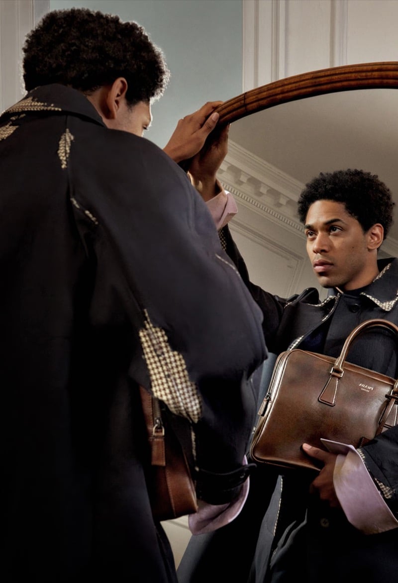 Kelvin Harrison Jr. adjusts a round wall mirror while holding a brown leather Prada briefcase, wearing a coat marked with a fractured tan print