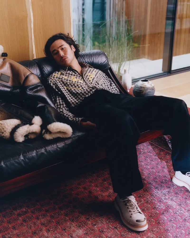 Nico Hiraga reclines on a black leather sofa wearing a plaid embroidered shirt, black trousers, and cream sneakers for Kenzo's fall-winter 2026 campaign.