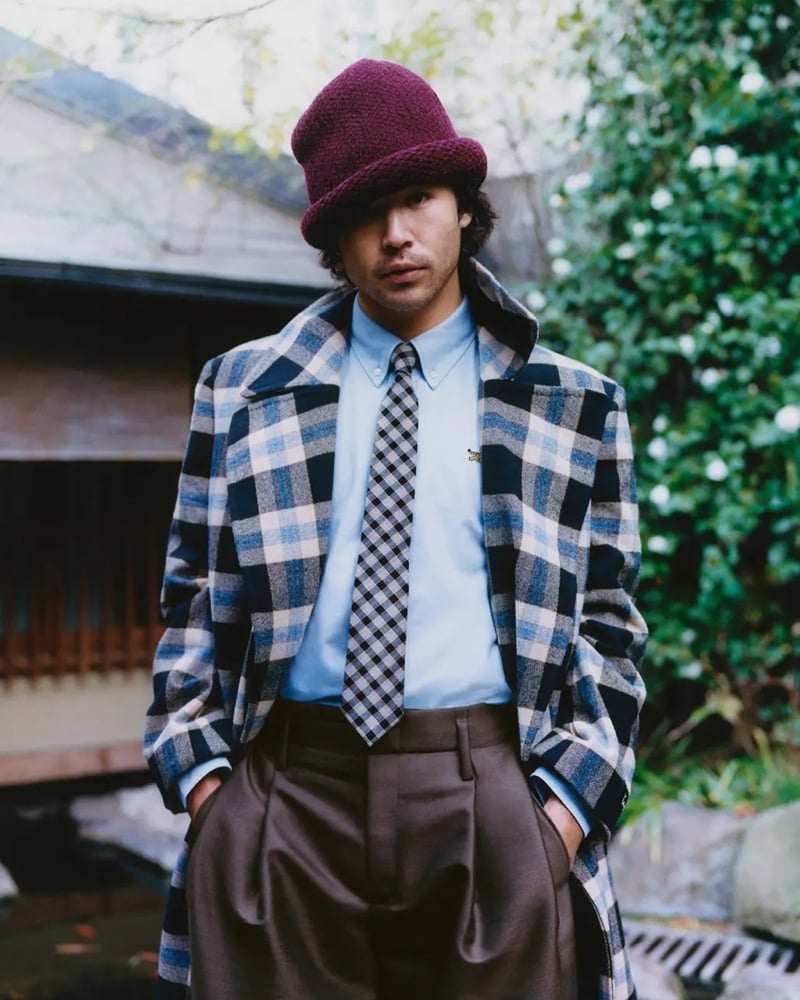 Nico Hiraga stands in a garden wearing a plaid coat, burgundy knit bucket hat, gingham tie, and brown pleated trousers for Kenzo's fall-winter 2026 campaign.