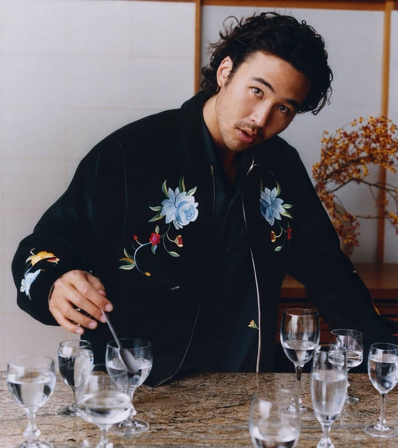 Nico Hiraga in a black floral-embroidered Kenzo bomber jacket sets a table lined with wine glasses inside a Japanese interior