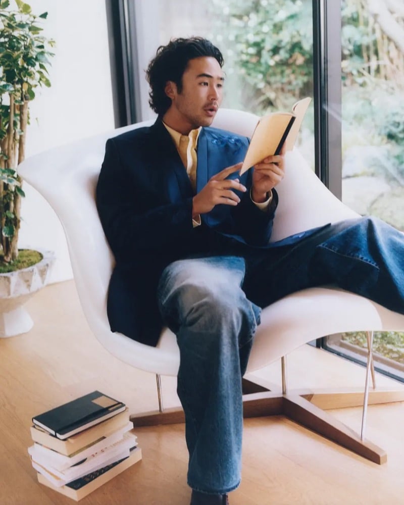 Nico Hiraga in a blue Kenzo blazer and wide-leg denim reads a book in a curved white lounge chair
