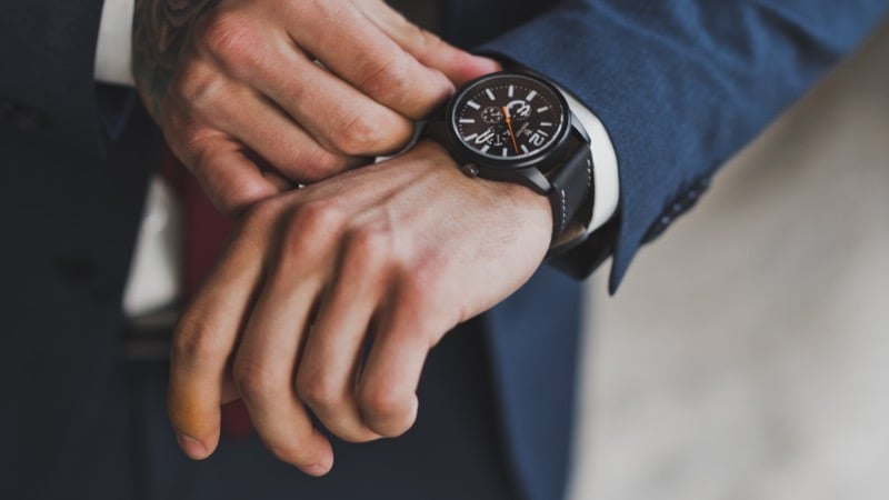A man in a navy suit checks a black chronograph watch on a leather strap