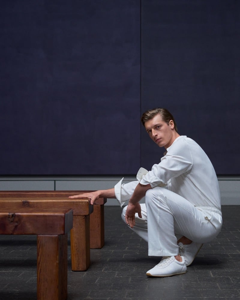 Colin Otto crouches beside a wooden bench in a white shirt and trousers inside a dimly lit room with dark paneled walls