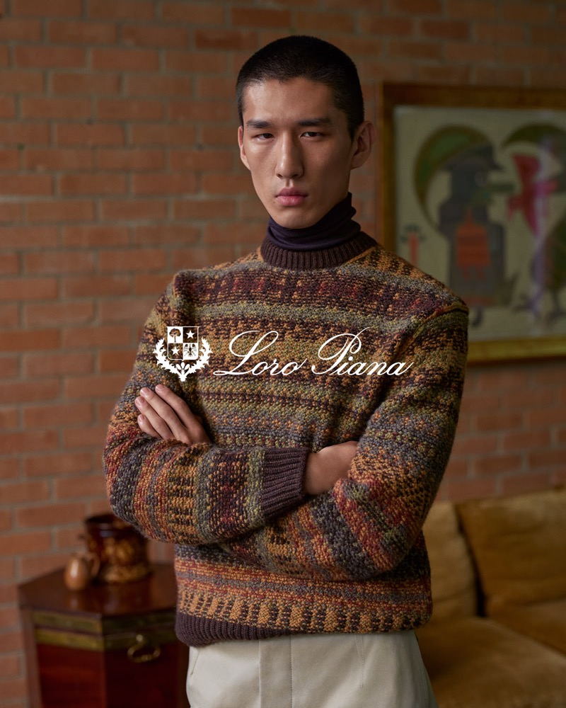 Long Li wears a multicolor Fair Isle sweater over a dark turtleneck, arms crossed in front of a brick wall hung with artwork