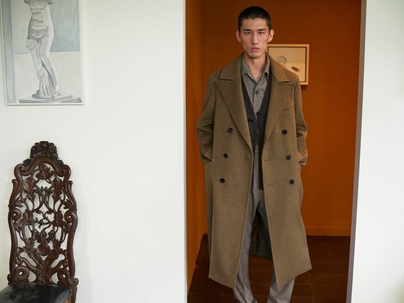 Long Li wears a camel double-breasted overcoat over a shirt and vest, standing in a doorway beside an ornate wooden chair