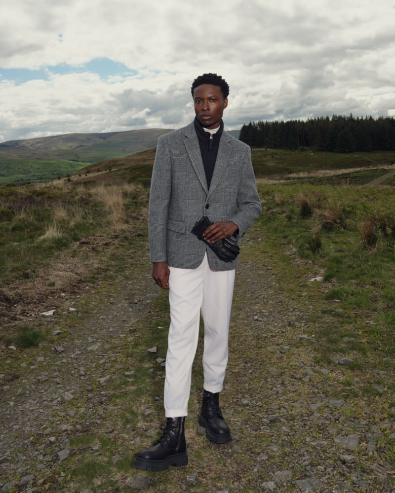 A man in a gray plaid blazer and dark quarter-zip sweater stands on a gravel path in the hills, wearing black lace-up boots and leather gloves