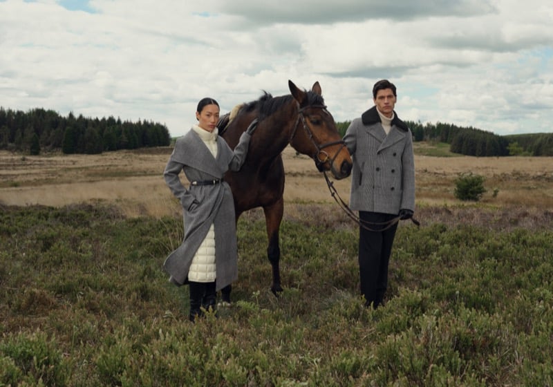 A woman in a belted gray plaid coat and a man in a gray plaid double-breasted coat with a shearling collar stand with a horse on a moor in the English countryside