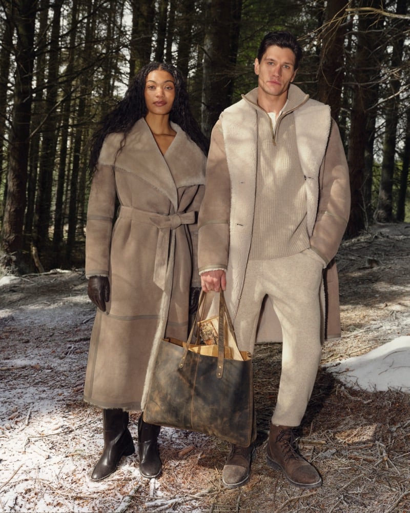 A woman in a belted shearling coat and a man in a shearling-trimmed jacket and ribbed sweater stand in a snow-dusted forest, a woven-front leather tote at her side