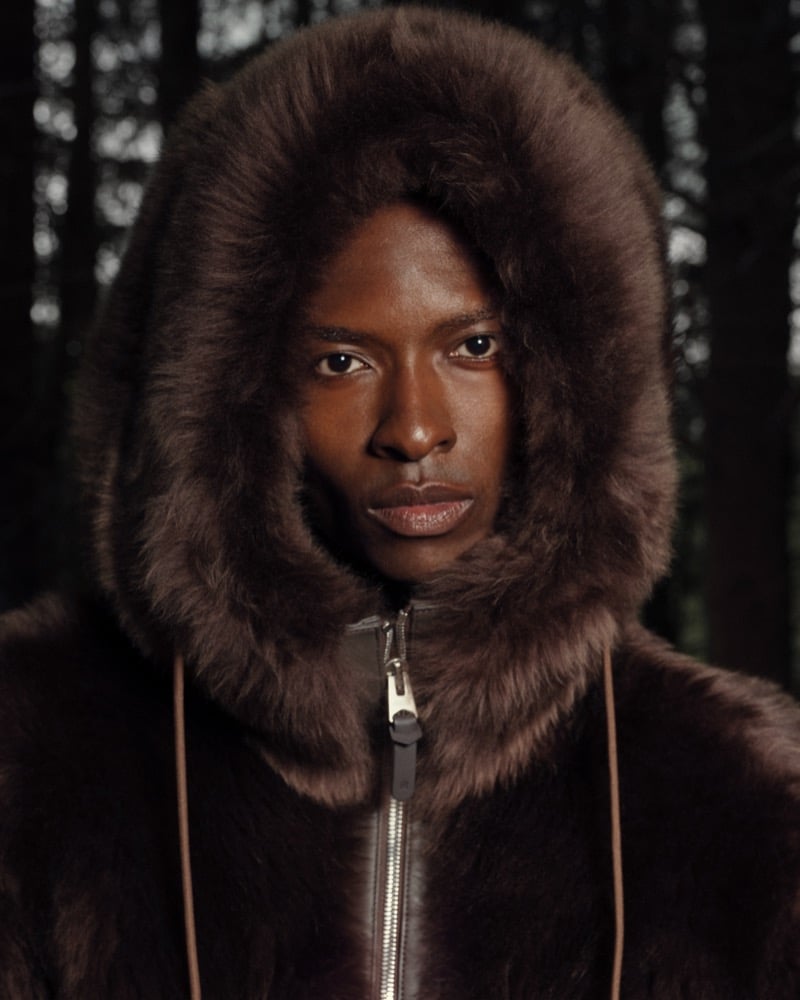 Close-up portrait of a man in a brown shearling-trimmed hooded jacket with the hood up, set against a wooded backdrop