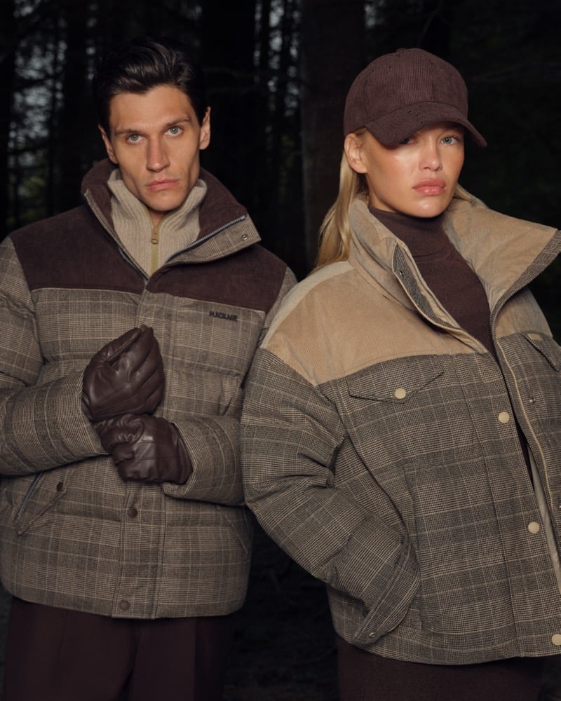 A man and a woman in checked puffer jackets with brown corduroy trim stand close together against a dark wooded backdrop, one wearing a corduroy cap