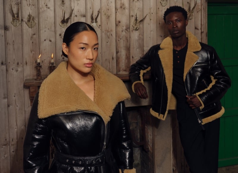 A woman and a man in black leather aviator jackets with tan shearling collars stand before a rustic wall decorated with antlers and lit candles