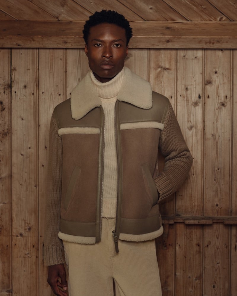 A man in a shearling-collared jacket and ribbed turtleneck leans against a wood-paneled wall in corduroy trousers