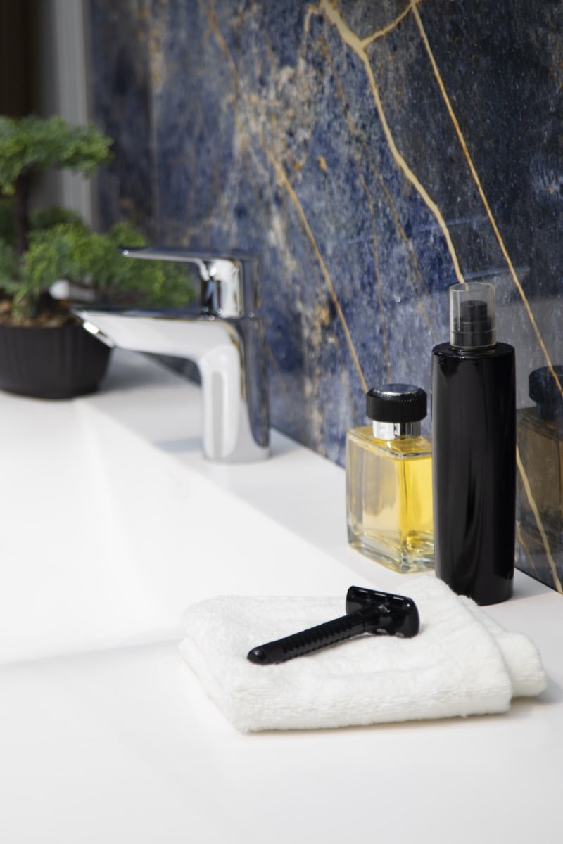 Overhead view of grooming products, a razor, and a folded towel styled on a bathroom counter with marble backsplash