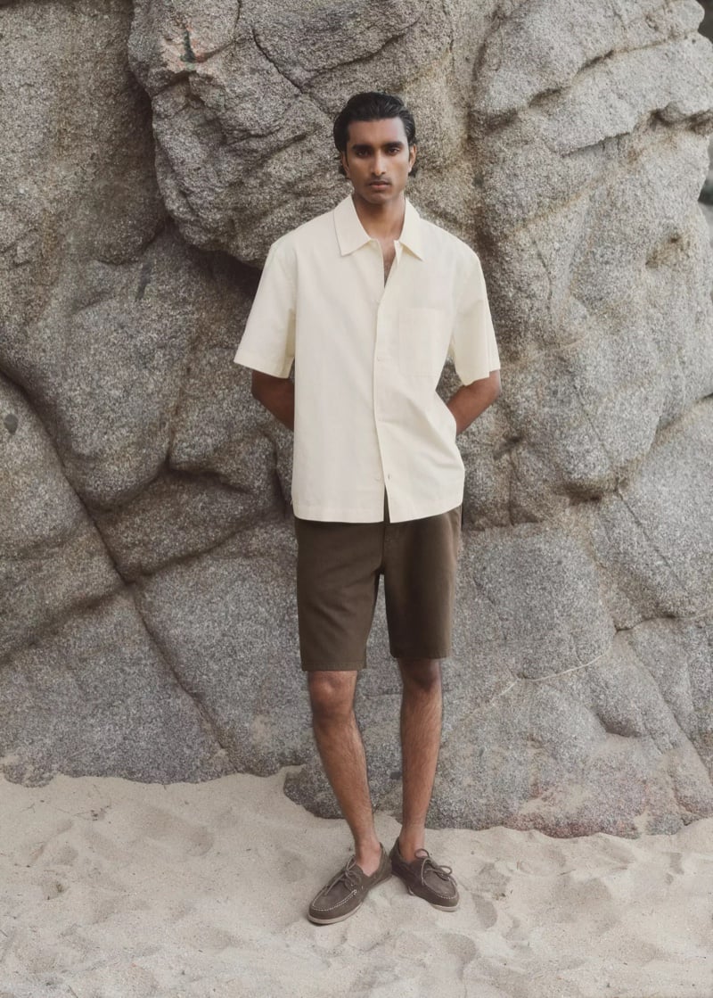 Jeenu Mahadevan in an ecru short-sleeve shirt and brown shorts with boat shoes against a rock formation