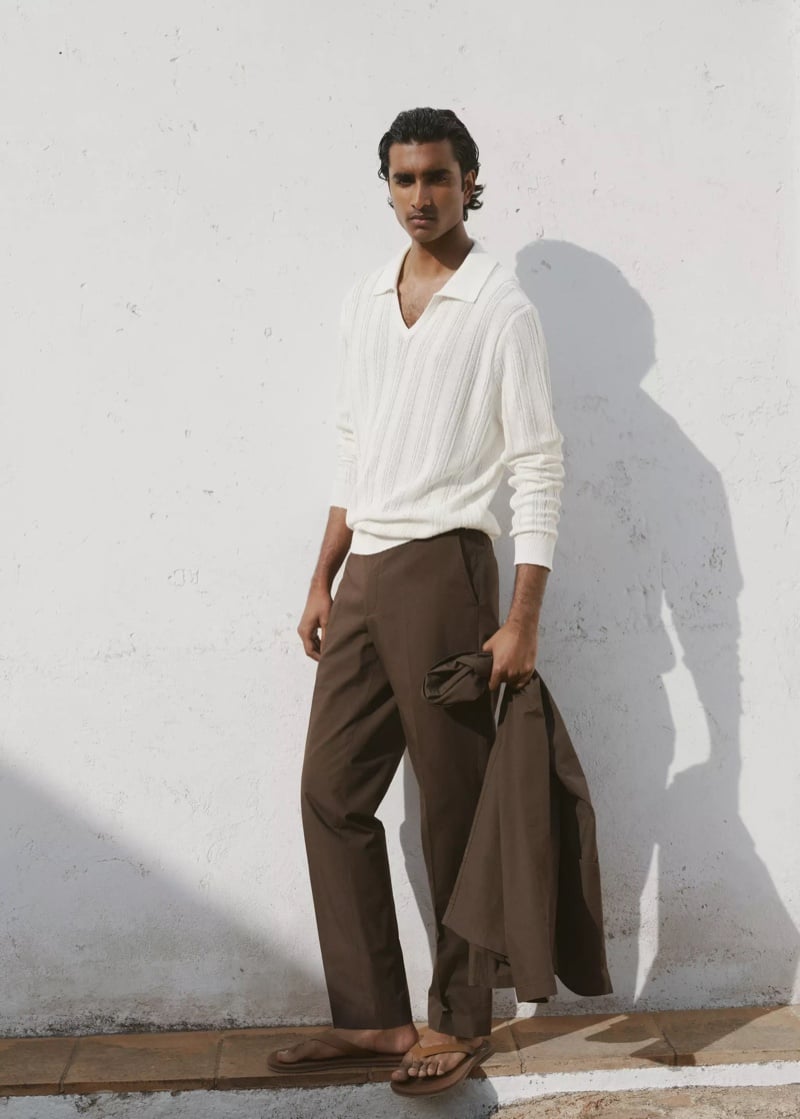 Jeenu Mahadevan in a white ribbed knit polo and brown trousers with a jacket draped over his arm
