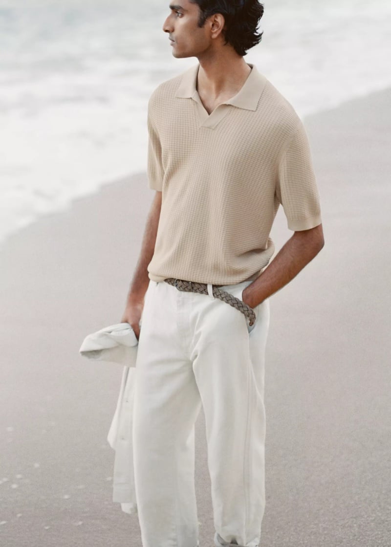 Jeenu Mahadevan in a beige knit polo walking along the beach holding a jacket