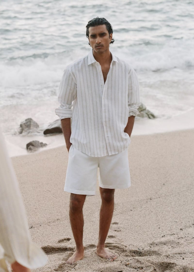Jeenu Mahadevan in a striped shirt standing barefoot on the beach near the waves
