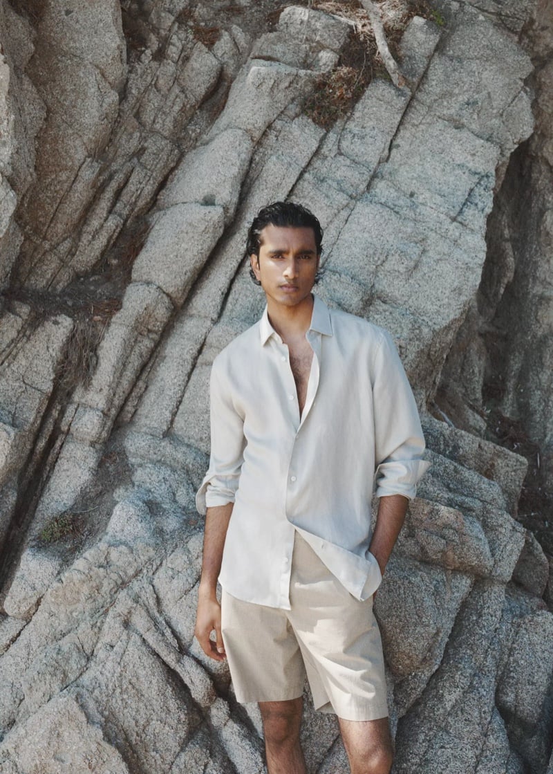 Jeenu Mahadevan in a half unbuttoned linen shirt and stone-colored shorts standing against a rock formation