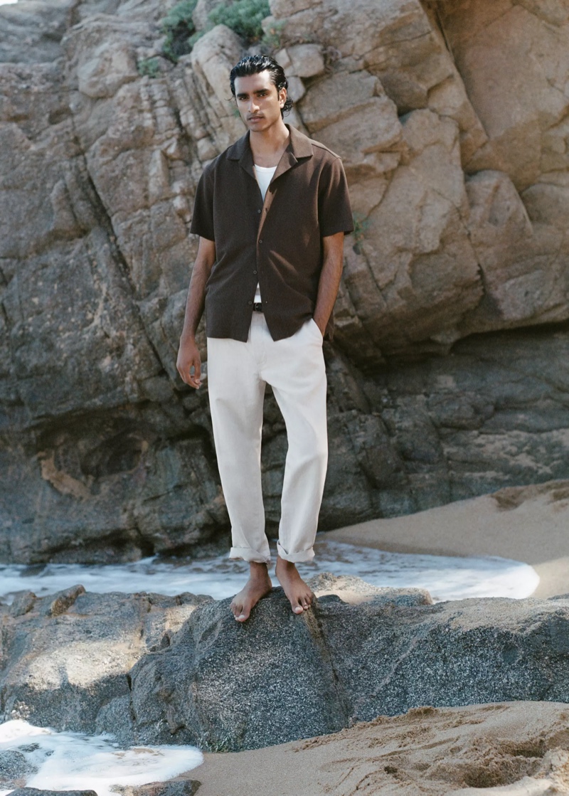 Jeenu Mahadevan in a dark brown knit camp-collar shirt standing on a rock at the beach