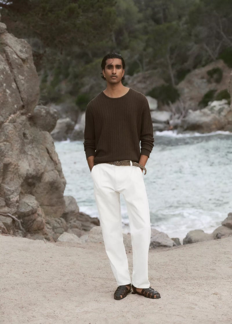 Jeenu Mahadevan in a brown ribbed sweater standing near a rocky, tree-lined cove