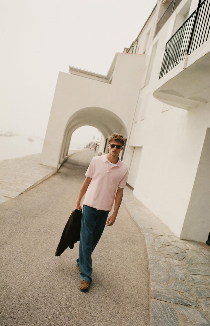 Vasko Luyckx walks along a coastal path in a pink polo shirt, dark wash denim trousers, and suede loafers, carrying a suede jacket over his arm past a white archway overlooking a hazy harbor