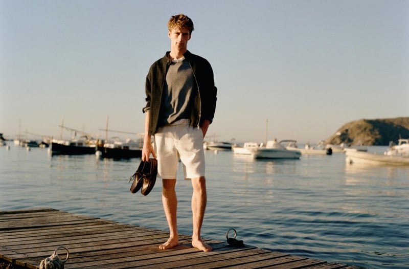 Vasko Luyckx stands barefoot on a wooden dock holding his shoes, wearing an olive zip jacket over a heather gray sweater and light-colored shorts, with harbor boats behind him