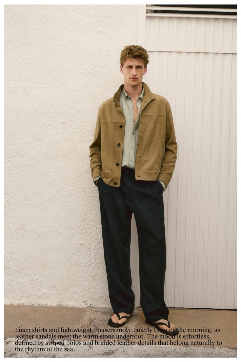 Vasko Luyckx wears a camel suede jacket over a sage green shirt and dark trousers, standing in sandals against a ridged white paneled wall