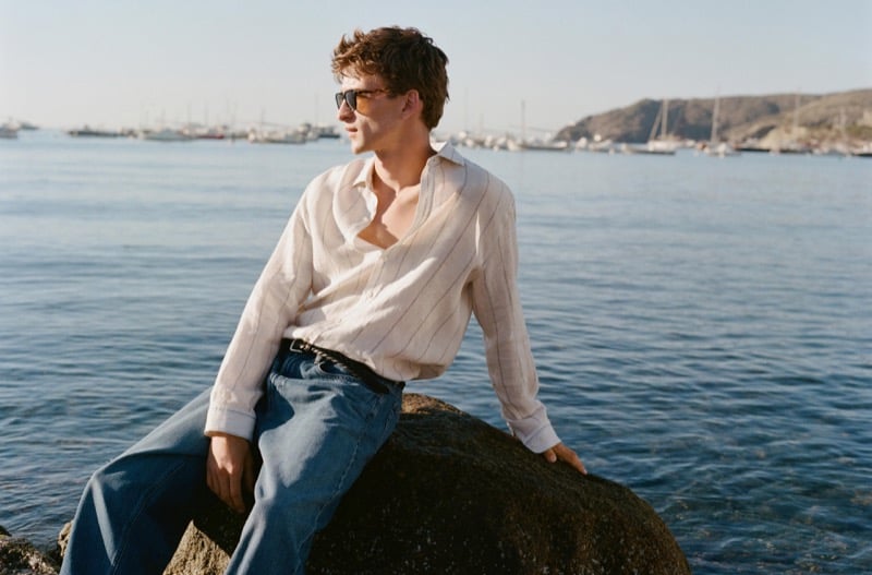 Vasko Luyckx sits on a rock by the harbor in an open pinstriped ivory linen shirt and dark wash denim, wearing sunglasses