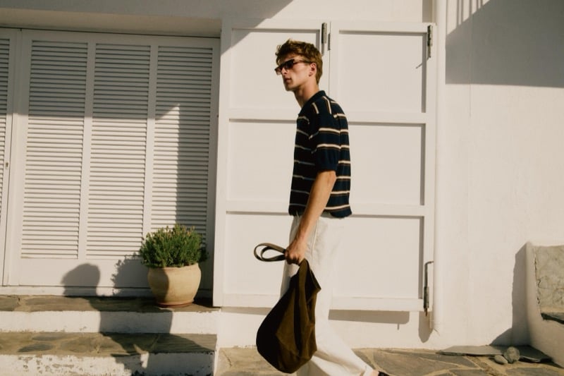 Vasko Luyckx walks past a white shuttered building carrying an olive suede tote bag, wearing a navy striped polo tucked into ivory trousers and sunglasses