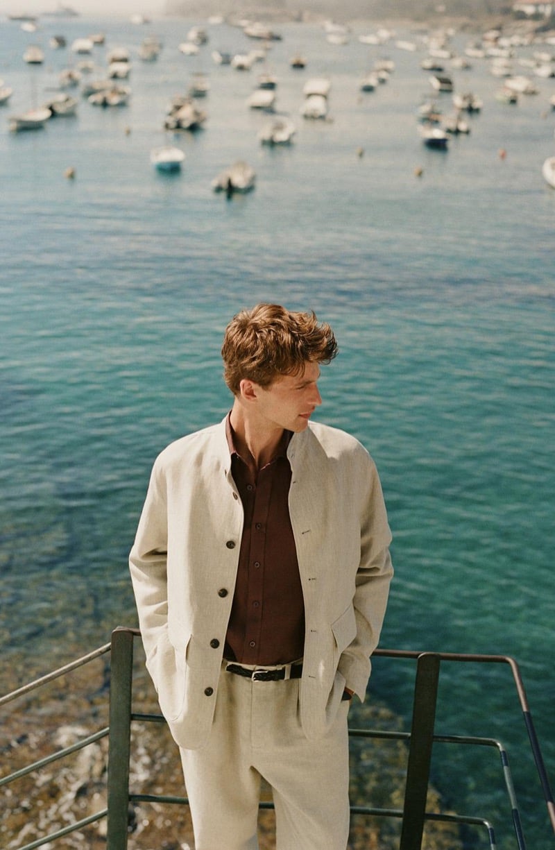 Vasko Luyckx wears an ivory suede jacket over a brown shirt and ivory trousers with a braided belt, standing at a railing overlooking a harbor full of boats