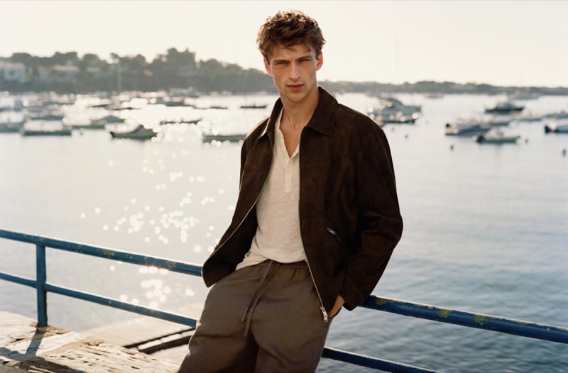 Vasko Luyckx wears a dark brown suede jacket over an ivory henley and olive drawstring trousers, leaning against a pier railing with boats glittering on the water behind him