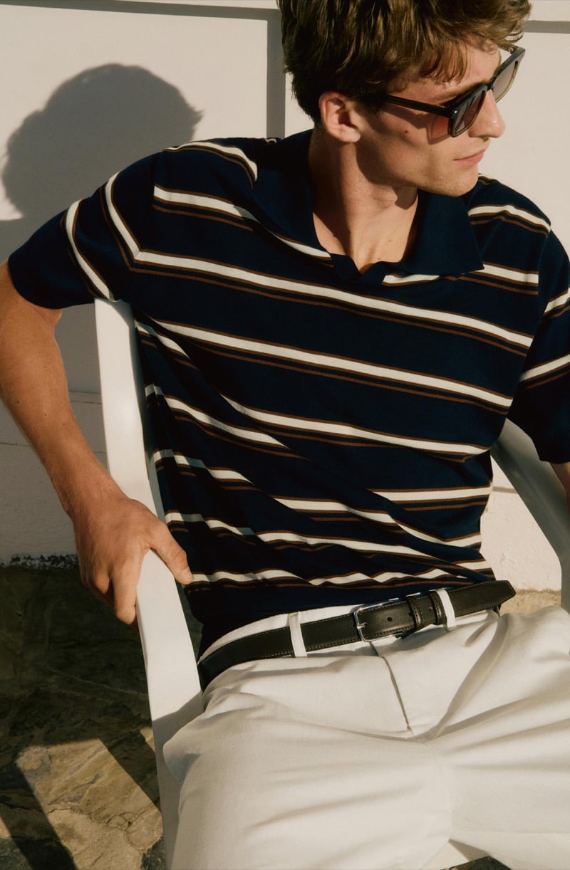 Close-up of Vasko Luyckx in a navy striped polo tucked into ivory trousers with a black belt, wearing sunglasses in a white chair