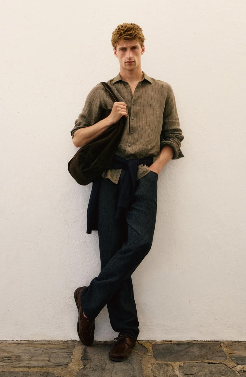 Vasko Luyckx wears a striped olive linen shirt with a dark suede jacket and sweater tied at his waist, dark trousers, and brown boat shoes, standing against a white wall