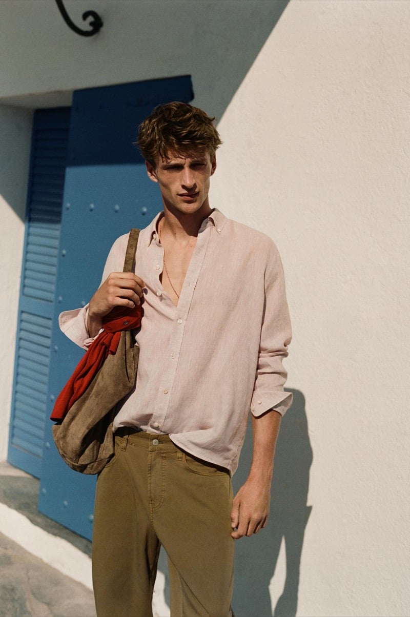 Vasko Luyckx wears an open blush pink linen shirt and olive trousers, carrying a suede bag with a red garment, standing before a blue shuttered door