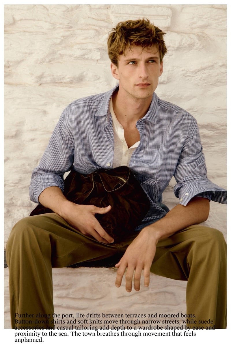 Graphic page of Vasko Luyckx in a blue gingham shirt over a white popover, holding a dark suede bag, wearing olive trousers against a textured white stone wall