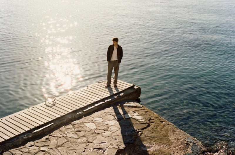 Distant aerial view of Vasko Luyckx standing at the end of a pier in a dark jacket and gray trousers, sunlight glittering on the water