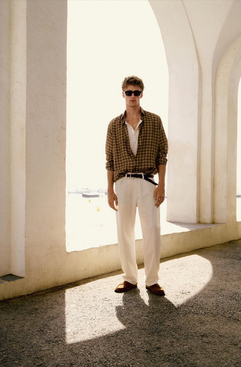 Vasko Luyckx wears a brown plaid shirt over a white popover with ivory trousers, a woven belt, and suede loafers, standing beneath a stone archway overlooking the harbor