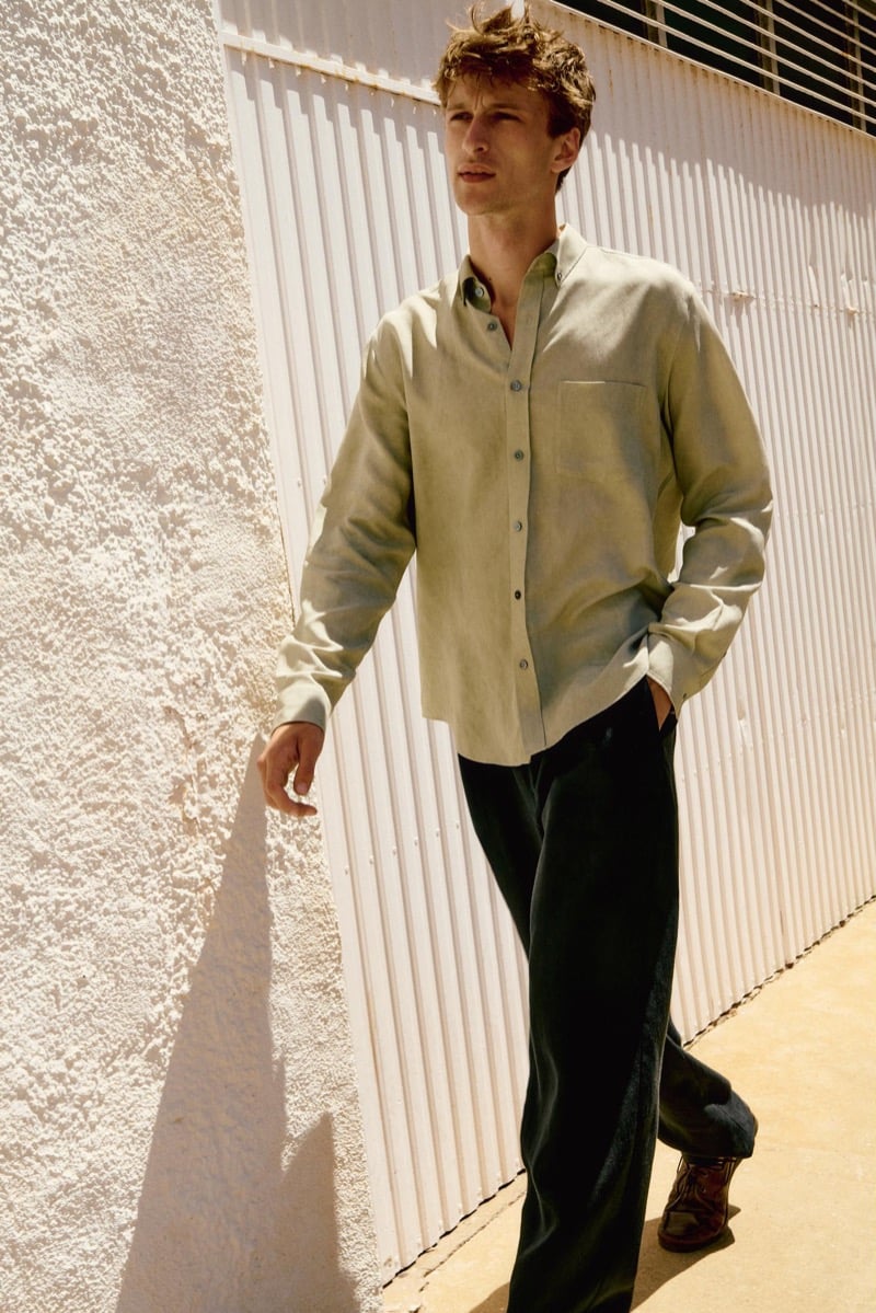 Vasko Luyckx wears a sage green button-down shirt and dark trousers, walking past a white corrugated panel wall in sunlight