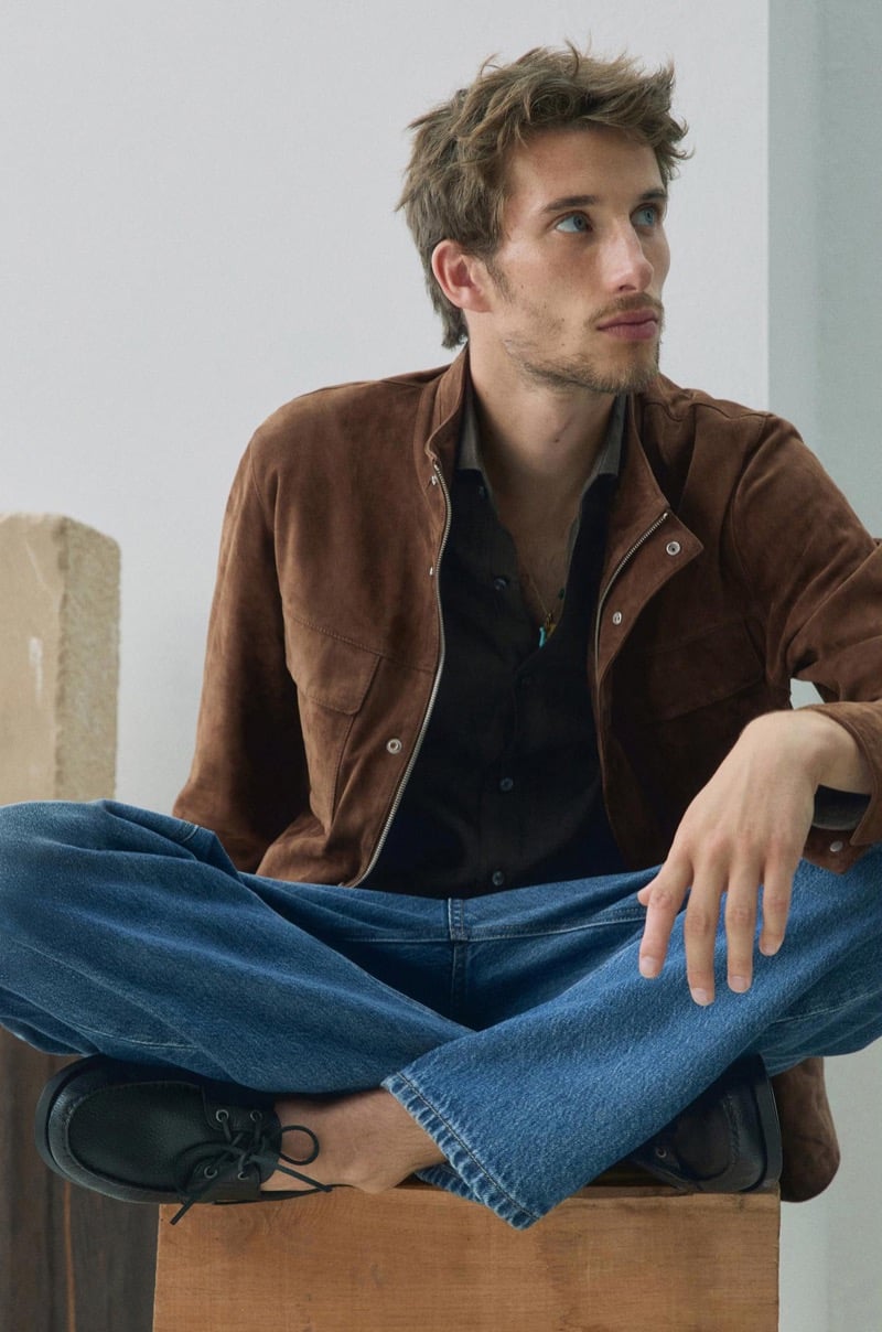 Aristide Deslandes sits cross-legged on a wood pedestal, wearing an open brown suede jacket over a black shirt with blue jeans and black loafers