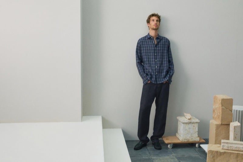 Aristide Deslandes stands against a wall in a blue plaid shirt and dark trousers with black loafers, looking upward