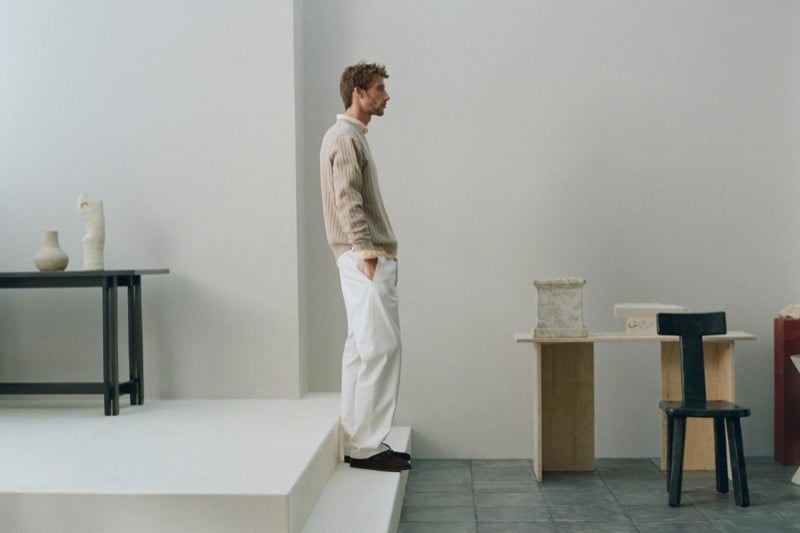 Aristide Deslandes stands in profile on a platform edge wearing an oatmeal ribbed turtleneck sweater with white trousers and dark loafers