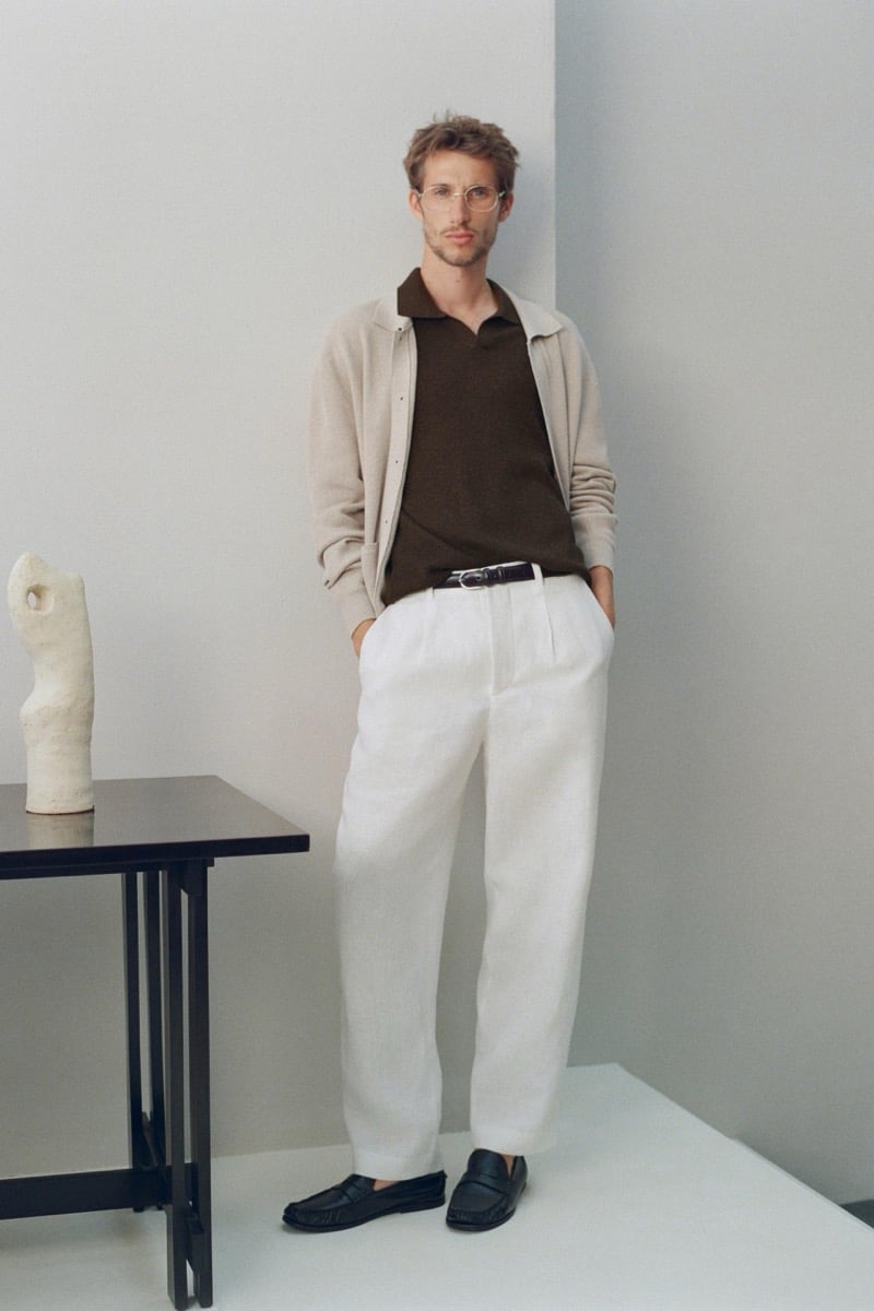 Aristide Deslandes stands in a beige-toned studio wearing an oatmeal cardigan over a brown polo, white pleated trousers, and black loafers