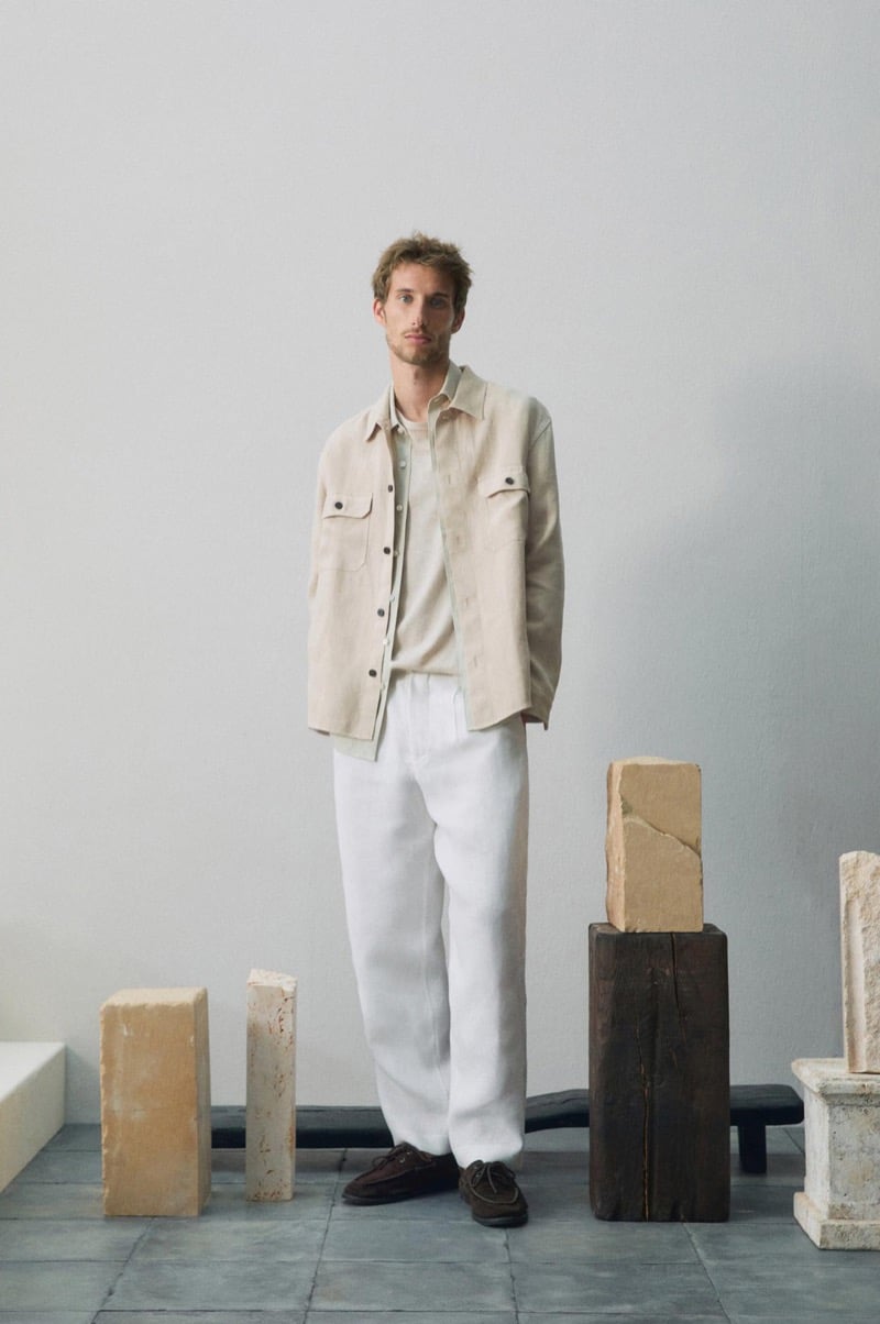 Aristide Deslandes stands among sculptural blocks wearing a beige shirt jacket over an oatmeal t-shirt, white trousers, and brown suede loafers