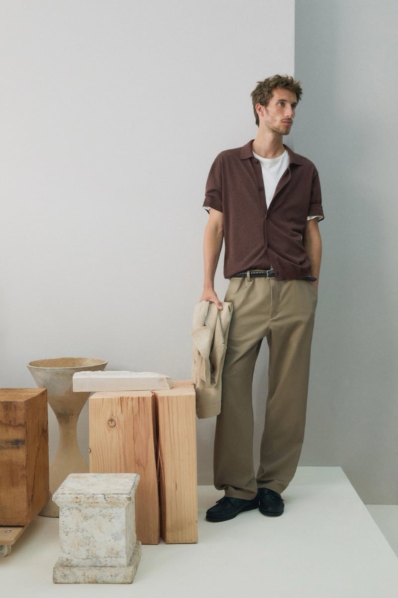 Aristide Deslandes stands holding a tan jacket, wearing a brown short-sleeve shirt over a white t-shirt with khaki trousers and black loafers