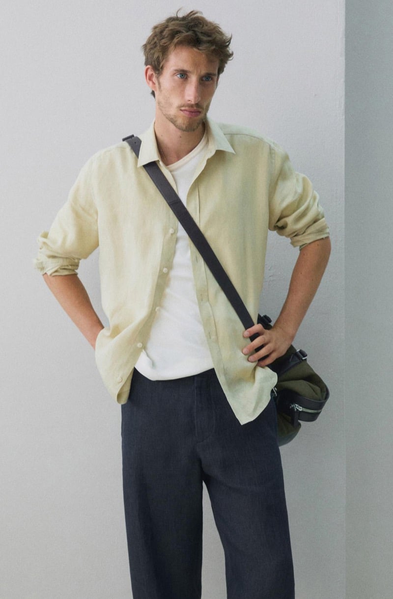 Aristide Deslandes stands with a hand on his hip, wearing a pale yellow linen shirt over a white t-shirt with a crossbody bag and dark trousers