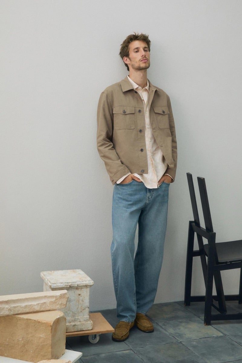 Aristide Deslandes stands against a wall wearing a tan jacket over a striped shirt with blue jeans and suede loafers