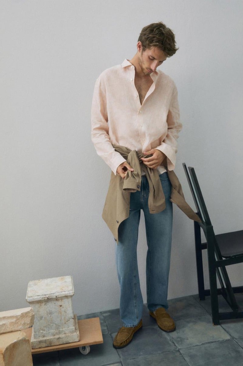 Aristide Deslandes stands with a tan jacket knotted at his waist over a pink linen shirt, blue jeans, and suede loafers