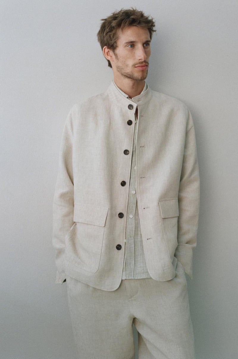 Aristide Deslandes stands against a wall wearing an oatmeal collarless linen jacket over a striped shirt with matching oatmeal trousers