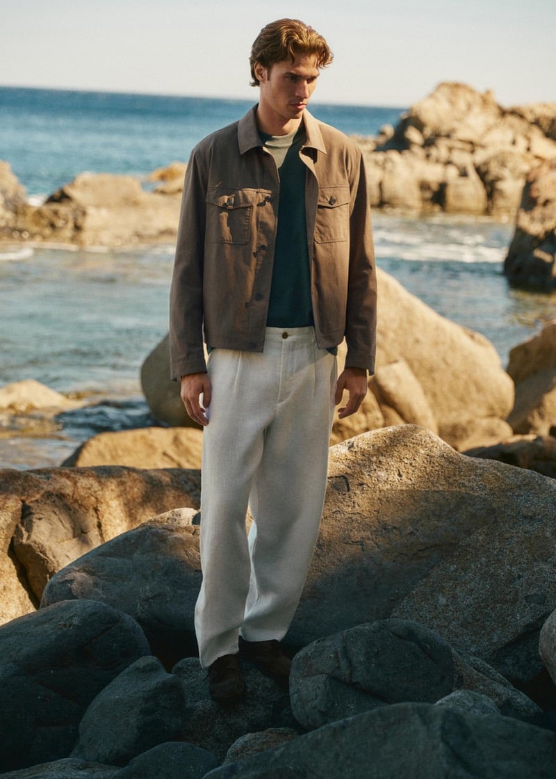 Callum Simpson stands on coastal rocks in a taupe field jacket over a teal T-shirt with ivory linen trousers, the sea and rocky shoreline stretching behind him